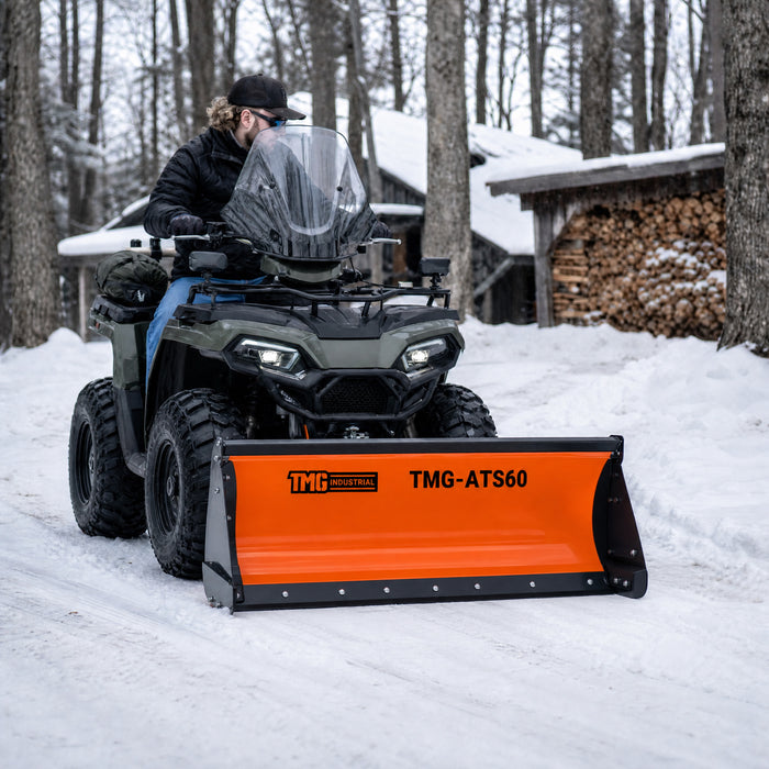 TMG-ATS60 snow bucket plow mounted on an ATV with a rider on a snow-covered path beside stacked firewood.