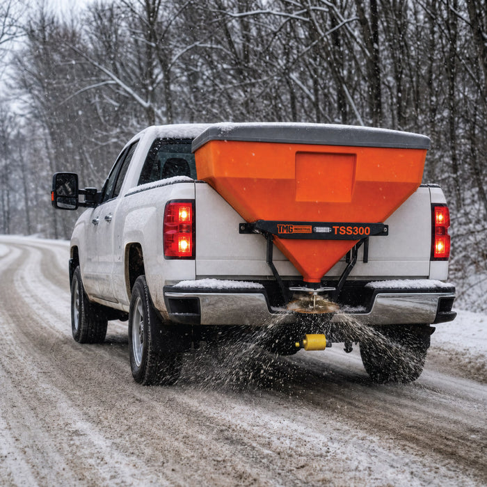 TMG-TSS300 tailgate salt spreader mounted on a white pickup truck spreading material across a snow-covered road.